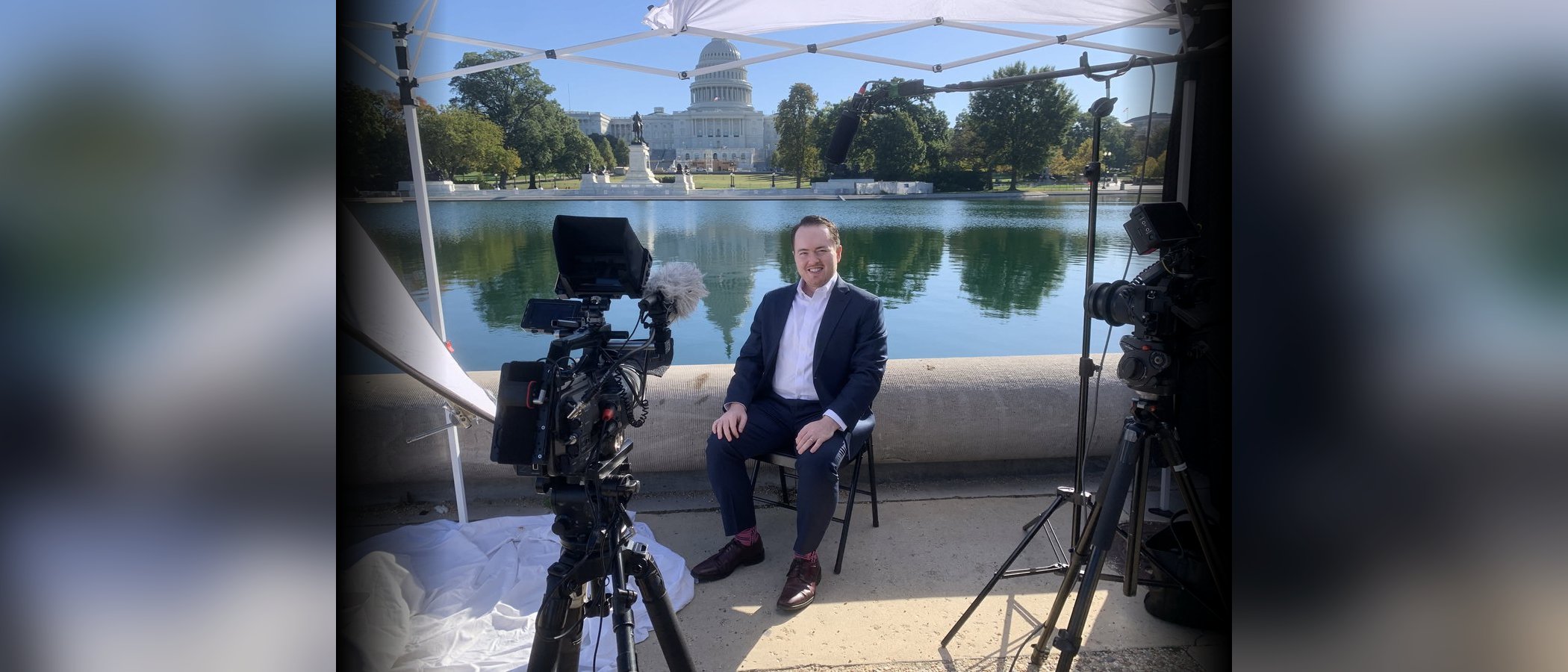 Seated for a broadcast interview beside the Capitol Reflecting Pool in Washington, cameras and lights around