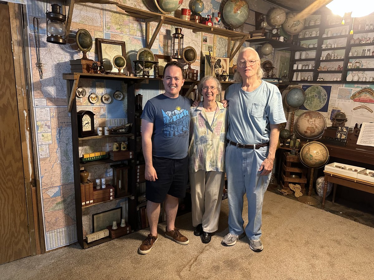 Standing with an older couple in a room lined floor-to-ceiling with maps, globes, and lanterns
