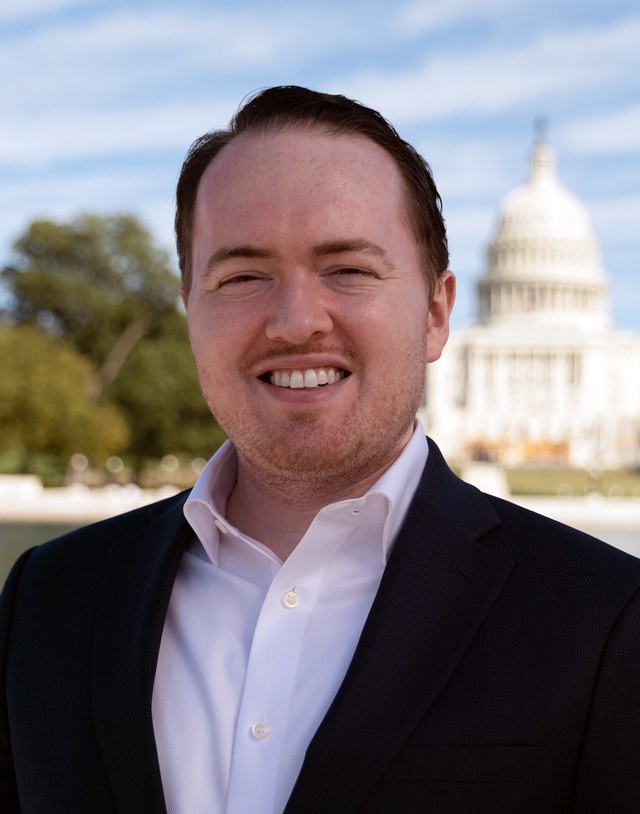 Professional portrait of Emerson T. Brooking in a dark suit and open-collared white shirt, photographed outdoors with the U.S. Capitol dome visible in the background