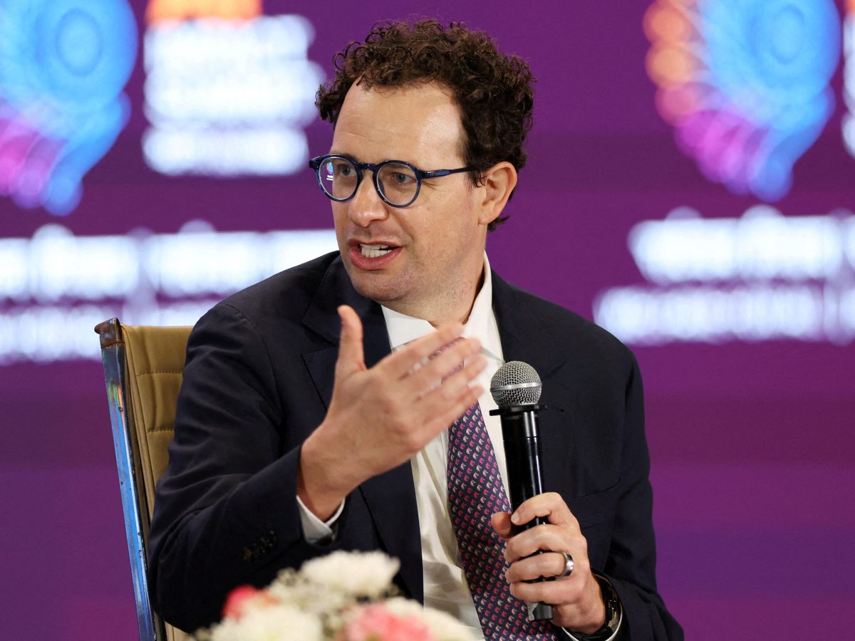 News photograph: Anthropic CEO Dario Amodei, microphone in hand, gesturing as he speaks from a chair onstage at the AI Impact Summit in New Delhi.