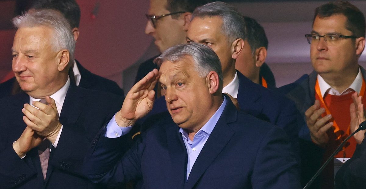 News photograph: Viktor Orban raising a hand to his temple amid a row of applauding party officials.