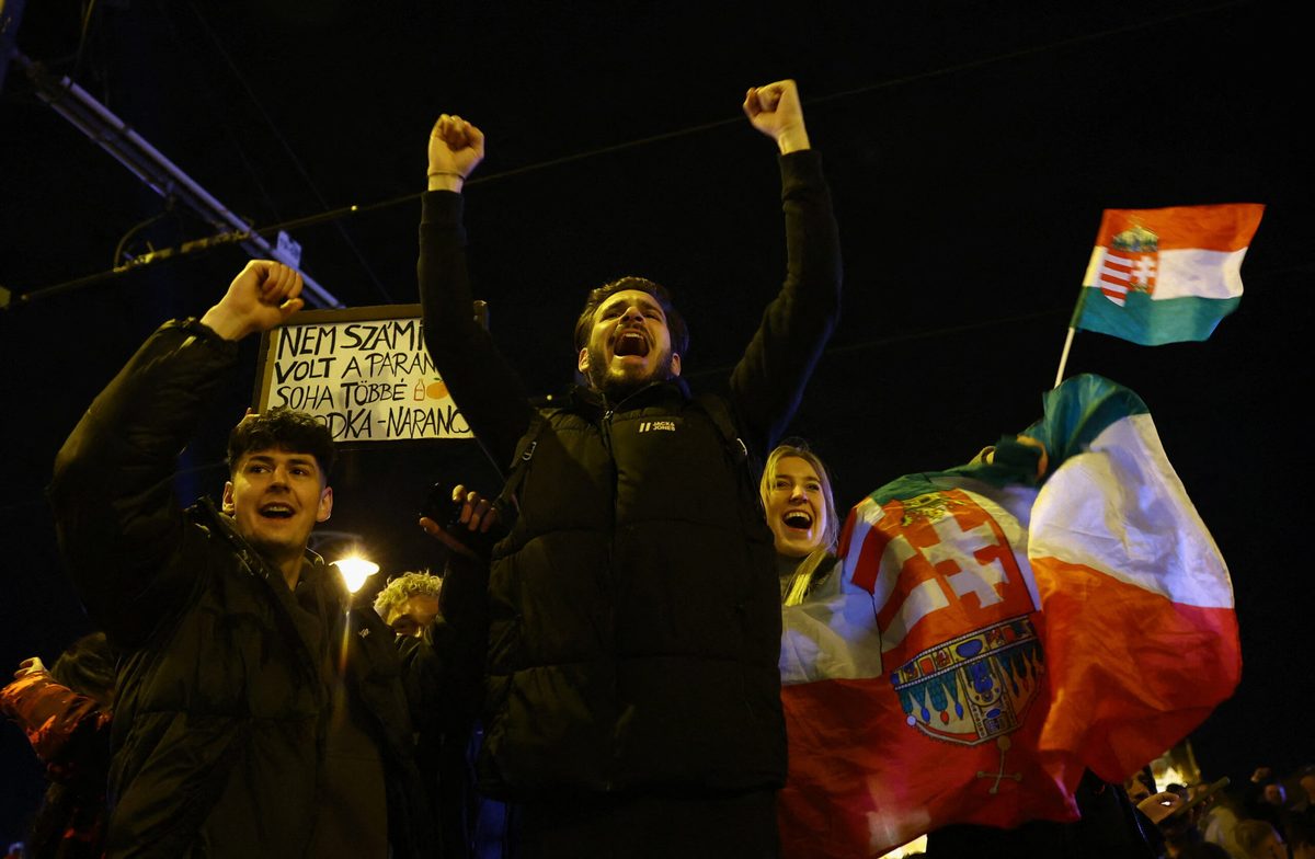 News photograph: a night-time crowd with fists raised, waving Hungarian tricolour flags and a hand-lettered placard.