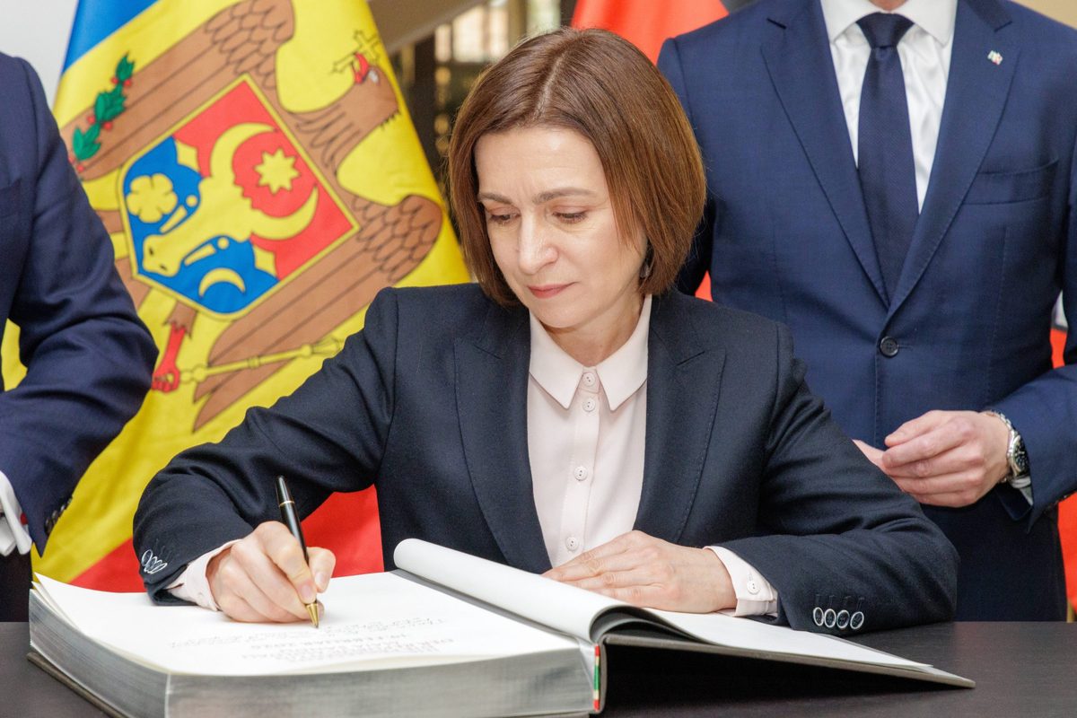 News photograph: Moldovan President Maia Sandu signing a bound register beside the Moldovan flag and coat of arms.