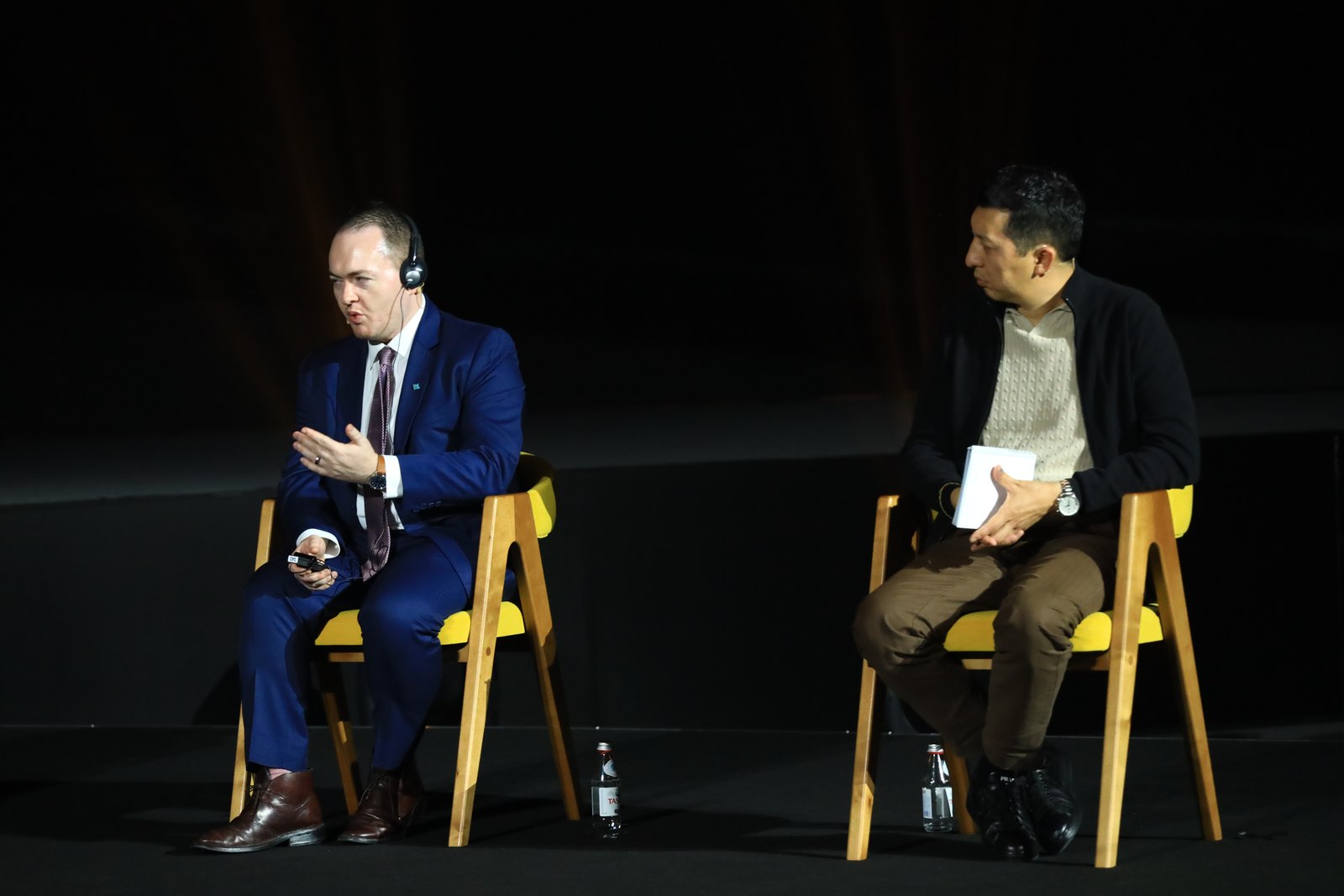 Seated in a navy suit and interpreter headset, mid-gesture, in conversation with a seated interviewer holding a notepad, on a dark QAZAQ Forum stage under a single spotlight