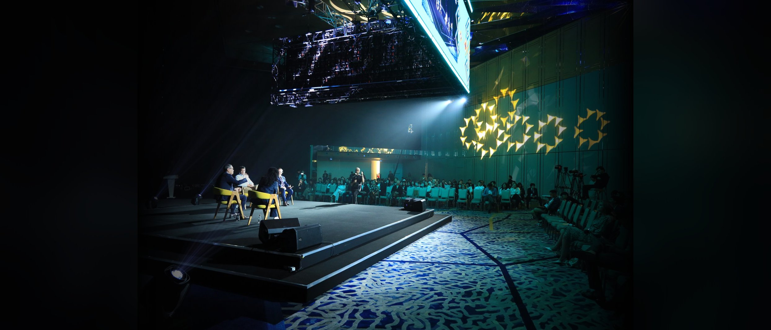Wide view of the four-person QAZAQ Forum panel seated on a dark stage under a geometric LED backdrop, Astana, May 2024
