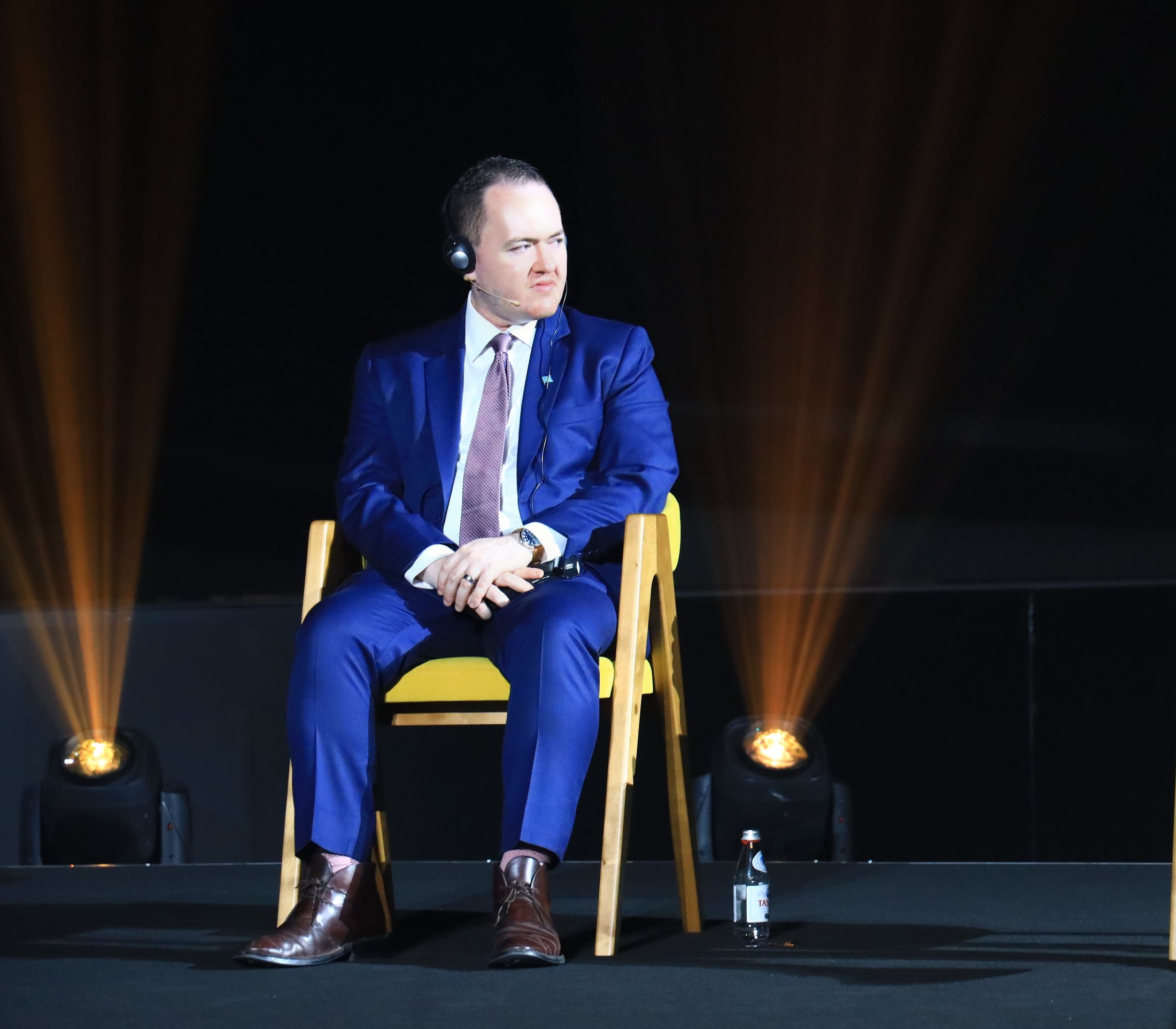 Seated on a dark theatrical stage under gold spotlight beams, in conversation with a host, wearing an interpreter headset