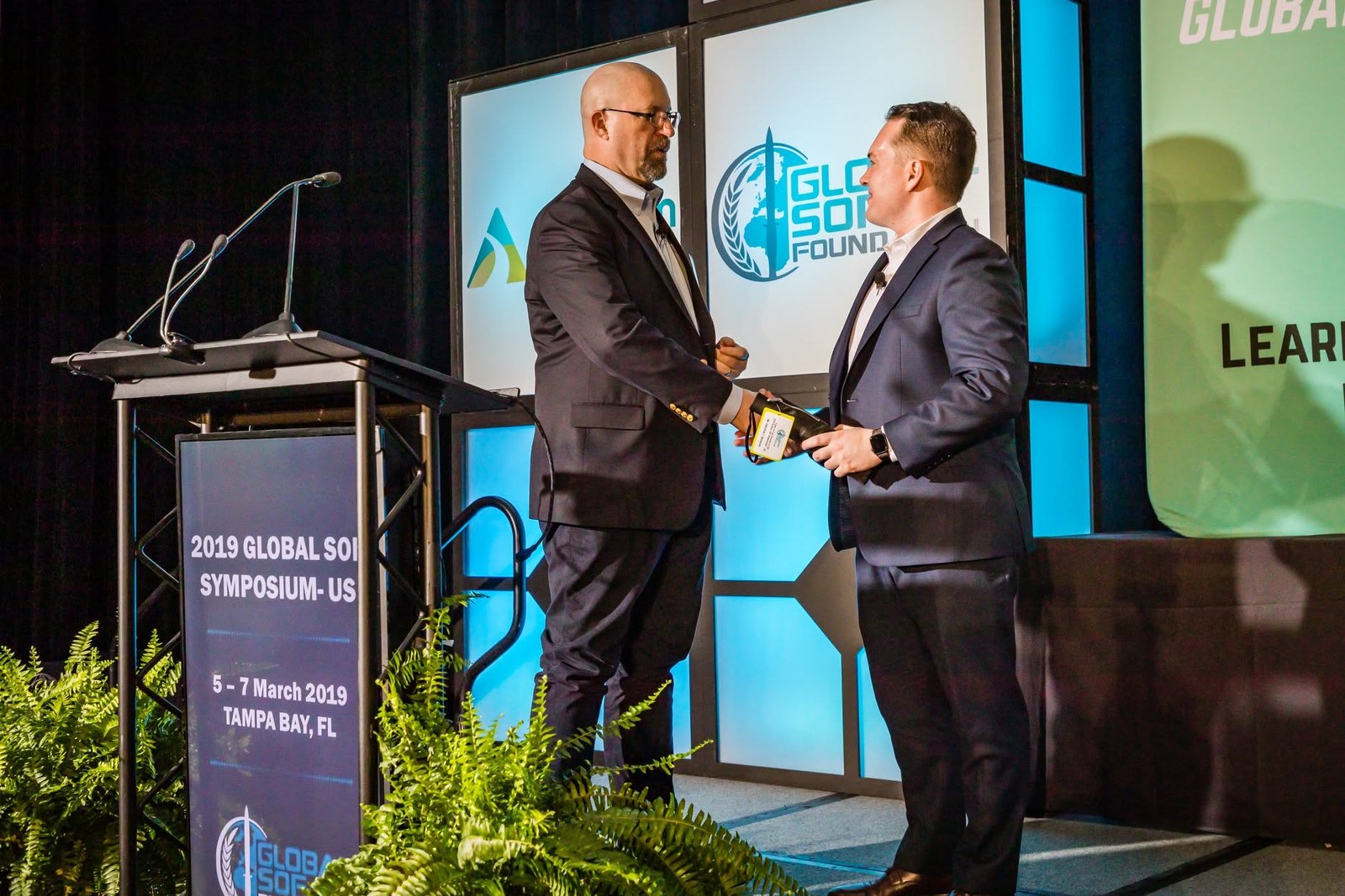 Shaking hands on stage at the 2019 Global SOF Symposium beside the lectern, a presenter's gift in hand
