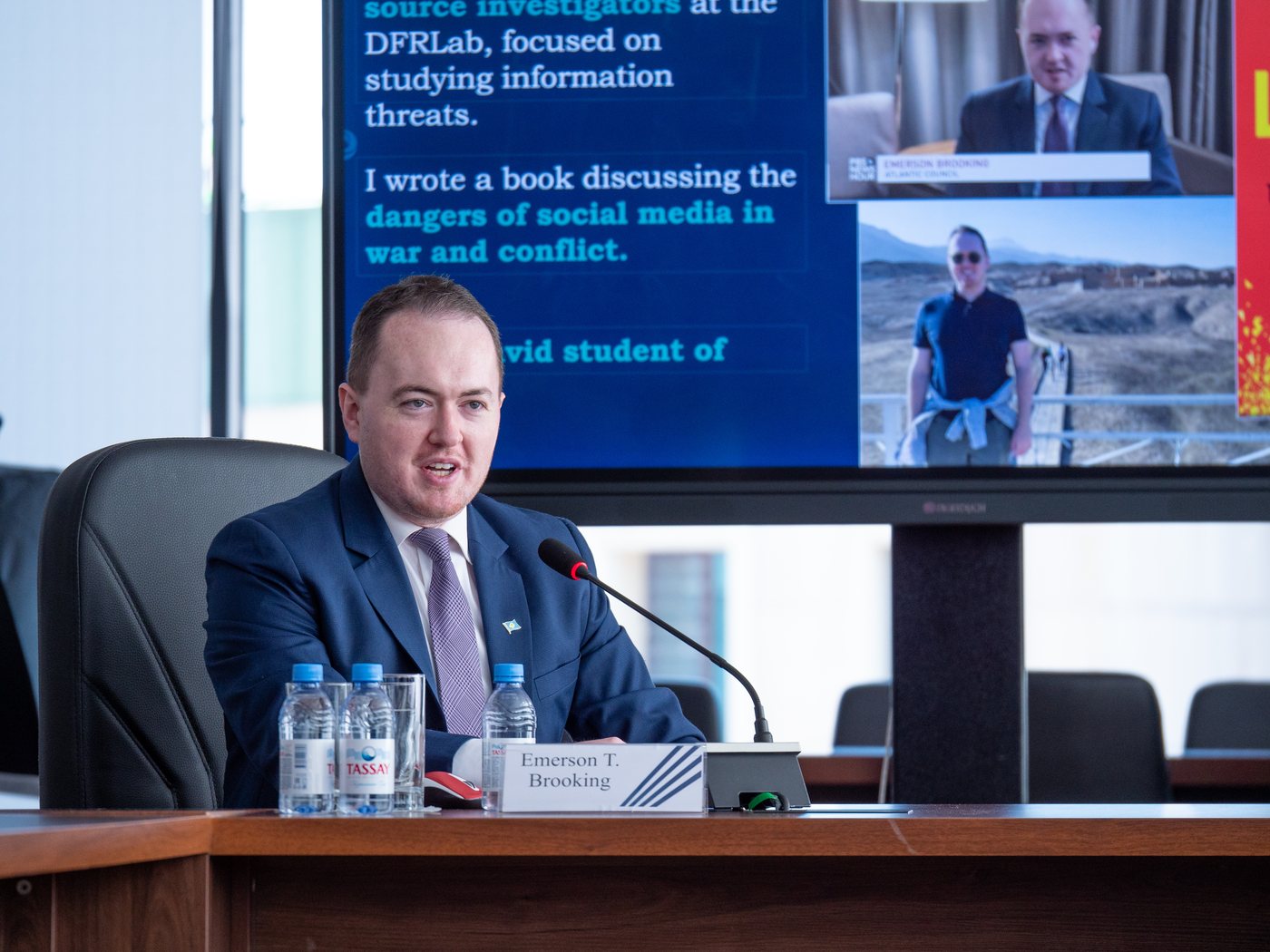 Speaking at a desk microphone beside a screen showing an About Me slide, Astana