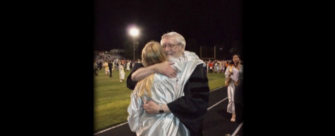 An older man in academic robes embracing a young woman in a graduation gown at a nighttime outdoor ceremony.